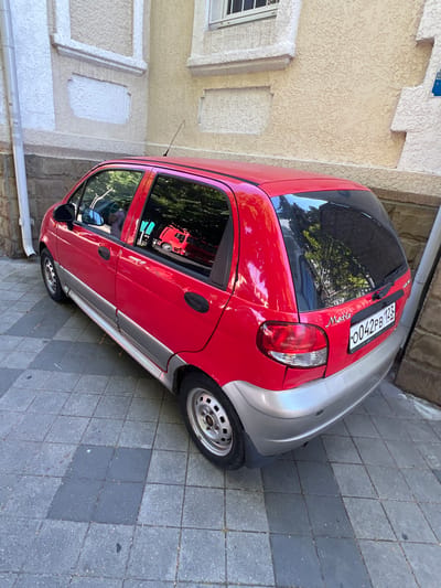 A red Daewoo Matiz hatchback is parked on a grey and dark grey tiled sidewalk next to a light-yellow textured building. The car's license plate reads "0042PB 123 RUS." The building has white decorative elements and a drainpipe runs down the left side. The car is viewed from a high angle, showing its rear and right side. The windows reflect trees and a red object, possibly another car. The scene appears to be an urban environment in Krasnodar, Russia.