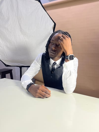This is a vertically oriented, medium shot photograph of a Black person dressed in formal wear. The person has braids and is looking directly at the camera with a serious expression. They are wearing a white dress shirt, a dark pinstripe vest, and a dark tie. A silver ring is visible on their right hand, which is resting on their forehead. On their left wrist, they are wearing multiple black beaded bracelets and a square smartwatch with a black band displaying "MITS" and "5:10". The person is leaning on a light-colored surface, possibly a table. To the left of the frame, a large white photography softbox is visible. The background consists of a plain, light brown wall and a white wall meeting at an angle. The lighting appears to be artificial, suggesting an indoor setting. There are no clear indicators of the time of day or weather conditions. The scene seems to be a professional portrait or studio setting.
