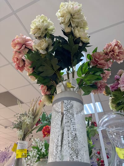 The scene depicts an indoor retail environment, likely a store, possibly located in the city of Klin, Russia, based on the Cyrillic text present on the price tags. The predominant subjects are floral arrangements, including artificial flowers in shades of white and pink, and bouquets that include artificial elements. A variety of vases and containers are also visible, including a white vase with a patterned design and a glass pitcher. Price tags are attached to some of the items, one showing a price of "790.00" and another featuring the text "140'00." The overall setting appears to be a retail display designed to showcase and sell floral decorations and vases. The background consists of a white, possibly tiled, ceiling.