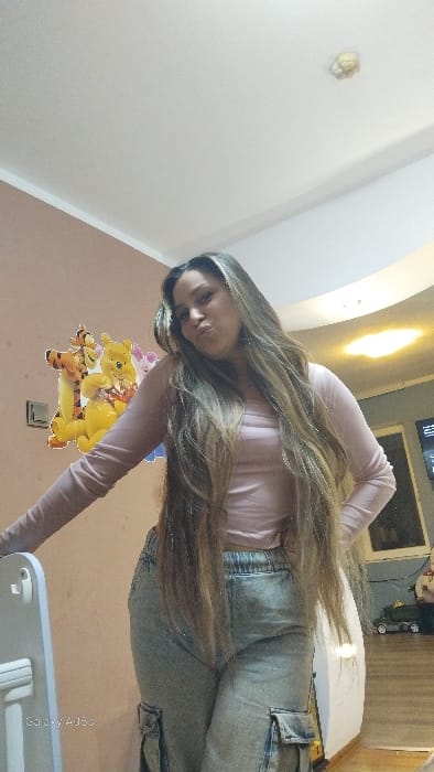 The image captures a female individual standing indoors, within a location specified as the city of Oreshene, Bulgaria. The subject has long, light brown hair with blonde highlights and is wearing a light pink long-sleeved top paired with light-wash denim cargo pants. She is facing the camera, with her head slightly tilted and lips pursed. Her left arm rests on a white, vertical object in the foreground, where a "Galaxy A05s" watermark is visible, while her right hand is positioned at her hip.
The room exhibits light peach-colored walls on the left and white walls on the right, with a white ceiling featuring a circular light fixture and a smaller, potentially functional ceiling device (e.g., smoke detector). The floor appears to be wooden. On the left wall, a decal depicting Winnie the Pooh, Tigger, and Piglet characters is affixed, positioned above a light switch. In the background to the right, a television screen displays content, and a dark-colored toy car is visible on the floor, adjacent to a white, curved architectural or furniture element.