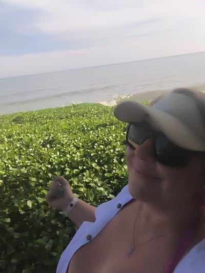 A woman wearing a visor and sunglasses is taking a selfie at the beach. She has on a white shirt and a pink bathing suit. The ocean is visible in the background. She is standing next to a dense patch of green vegetation. The location is Phu Thuy Ward, Vietnam.