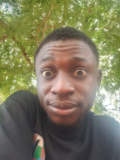 A medium-close-up shot of a Black man with short, dark hair, wearing a black shirt. The man is looking directly at the camera with wide eyes. The background shows green foliage, suggesting an outdoor setting. The image's upward angle implies the photo was taken from a low position. There is no verifiable evidence to place the photo within Adankolo, Nigeria.
