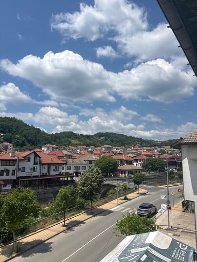 The image depicts a view of the city of Zlatograd, Bulgaria, on a sunny day with scattered white clouds in a blue sky. The scene looks down onto a street with a grey asphalt surface and a yellow-tiled sidewalk. On the left side of the street, a low stone wall with a metal railing separates the sidewalk from a small, dry riverbed or canal. Behind the wall, several trees with lush green foliage are visible.
Across the street, a row of buildings with red-tiled roofs lines the embankment. These buildings appear to be a mix of residential and commercial properties, with some displaying signs in Cyrillic. One building has a sign that reads "КЪЩА ЗА ГОСТИ ТОНИ" (Guest House Toni). Further back, the town extends up a verdant hillside covered in dense green trees.
A light-colored car, possibly a hatchback, is driving on the street, heading away from the camera and towards a bridge in the distance. The bridge appears to be an old stone structure. There are pedestrian crossings marked on the road. On the right side of the street, the edge of another building with a white facade and a grey tiled roof is visible, along with a portion of what looks like a canopy or awning with text and graphics. A lamppost stands near the curb. The overall impression is of a charming, historic town nestled within a hilly, green landscape.