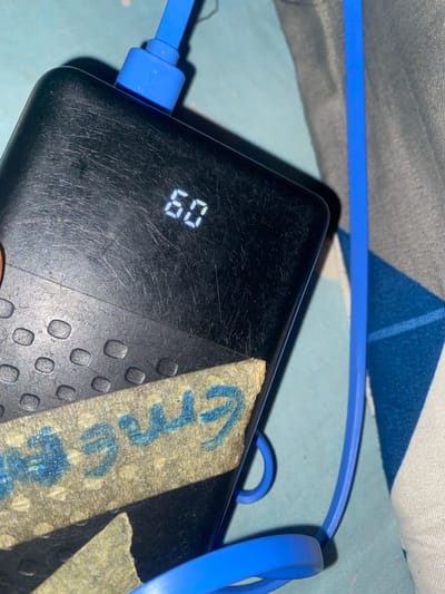 The image shows a close-up of a black power bank that is currently charging. A blue charging cable is plugged into the power bank. The power bank has a digital display showing "60", likely indicating the battery percentage. There are several small, circular raised dots on the surface of the power bank. A strip of tan-colored tape with blue writing is affixed to the power bank. The blue writing is partially obscured but appears to be a name or word, with letters like "Gree" visible. The power bank is placed on a surface that has a blue and white checkered pattern, possibly a cloth or bedding. The lighting suggests it is indoors, with no strong indications of the time of day or weather conditions. The overall impression is a functional object in a domestic setting.