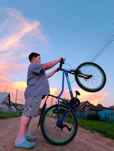 In Зипуново, Russia, a young boy stands on a dirt road, holding the handlebars of a blue bicycle upside down. The sky is a vibrant mix of blue and pink clouds, suggesting sunset. The boy wears a gray t-shirt and gray shorts, with light blue clogs and white socks. The bicycle has green rims on its wheels. In the background, there are wooden houses, a blue fence, and green grass. Electrical wires are visible against the sky. The scene appears to be in a rural setting.
