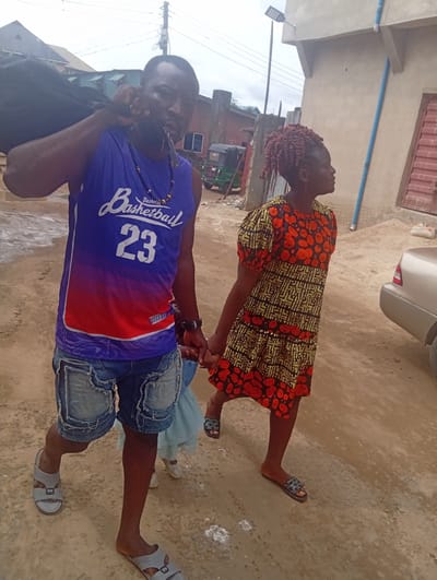 Two adults walk hand-in-hand on a dirt road in Umuode, Nigeria. The man wears a blue and red basketball jersey with the number 23, jean shorts, and grey sandals. He carries a black object over his left shoulder and has a key in his mouth. The woman is in a short-sleeved dress with orange and black patterns and slides. A small child in a light-colored dress is partially visible between them. In the background are buildings of different styles, including a light grey building with a water pipe and a parked car. An auto rickshaw is visible in the distance. The sky is overcast.