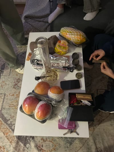 The scene is an indoor table, likely in a living room, located at coordinates 40.951, -4.119, which places it in the city of Segovia, Spain. A white table with a white tablecloth is centered in the frame, covered in various objects. A large glass bong sits on the table with a bowl on top. Four mangos are packaged in clear plastic containers, each with a net-like covering. The mangos are arranged to the left of the bong and the bowl, A yellow package labeled "Mission Street Tacos" sits to the right of the bong. Two grinders with dark grey metal lids sit on the table to the right of the taco package. A black box with a wooden lid is to the right of the mangos. The box contains a red pouch, green items, and a white piece of paper. A black cell phone sits on top of the box. A purple pouch is below the box. Two pairs of keys sit to the right of the pouch. A small, clear bag containing green vegetation is below the mangos. A dark brown blanket is on the floor behind the table. The image is taken from a slightly high angle, looking down on the table. Two people, likely in their teens or early twenties, are partially visible in the image. One is wearing blue jeans and a black shirt. The other is wearing green cargo pants and a black shirt. Both are sitting on the floor with their feet extended towards the table. The person in green is wearing white sneakers and their right foot is on the table. The person in blue jeans is wearing white socks with dark shoes. Their right hand is on the table and their left hand is on the floor. The image is likely taken by someone standing in the room, looking down on the table. The image is of a gathering of friends enjoying a relaxed moment together, likely partaking in recreational activities.
