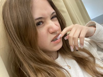 A young female with long, light brown hair is depicted in a close-up, indoor shot within Raanana, Israel. Her face is centered, displaying a scrunched expression with a wrinkled nose and forehead, eyes slightly narrowed. She is wearing clear orthodontic braces on her upper teeth, visible as her upper lip is slightly drawn up. Her right hand is positioned with the index finger touching her upper lip. The fingers feature long, clear, rectangular acrylic nails; the nail on her ring finger has a small black and orange design. She is dressed in a cream-colored knit sweater, and a thin gold-colored necklace with a small pendant is visible. A wrist device, possibly a smartwatch, is partially visible on her left wrist beneath the sweater cuff. The background consists of light beige fabric, possibly a curtain or drapery, and a plain light-colored wall.