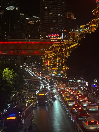 The media file depicts a bustling city scene at night, likely in Yuzhong District, China, characterized by vibrant lights and heavy traffic. The perspective is from an elevated viewpoint, looking down upon a densely populated urban environment.
The primary subjects are a multitude of cars and buses lining a road, many with their headlights and taillights illuminated, indicating a significant traffic jam. The vehicles are closely packed, suggesting congestion. Numerous people are visible, either walking along the streets, gathered on sidewalks, or possibly in viewing areas, their presence contributing to the lively atmosphere.
The scene is set against a backdrop of towering buildings, many adorned with bright, decorative lights, showcasing a mix of modern architecture and traditional-inspired designs, particularly on what appears to be a pedestrian street or market area. The overall ambiance is one of activity and festivity, with the abundance of lights suggesting a special occasion or simply the energetic nightlife of a major city.
Notable details include the time of day, which is clearly night, as indicated by the darkness and the prevalence of artificial lighting. There are no obvious cues about the weather, but the scene suggests a clear night. The abundance of lights, the crowds, and the traffic point towards a lively urban environment.
Visible text includes Chinese characters on signs and on the vehicles. For instance, some vehicles have illuminated text in Chinese, such as "0401" on one of the buses. The architectural elements also display illuminated signage, though specific characters are not clearly legible in all instances. The overall impression is a dynamic and vibrant urban landscape at night, filled with people and vehicles.