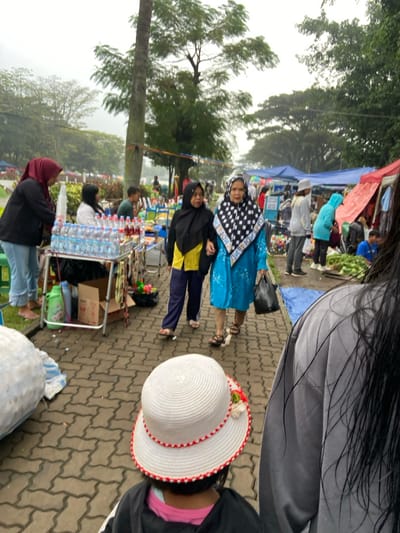 The image shows a street market in Bandung, Indonesia, located at coordinates -6.896 latitude and 107.619 longitude. The market is held on a paved street with brick-like blocks, lined with trees and vendors. A young girl wearing a white sun hat is in the foreground. Two women are walking towards the camera, one wearing a blue patterned headscarf, a blue patterned long dress, and a black plastic bag, and the other woman wearing a black headscarf, a yellow long-sleeved shirt, and black pants. There are other people walking around the market and behind the two women in the foreground. Vendors are seen setting up their stalls along the side of the street. The scene appears to be a typical street market in a bustling city. The weather is cloudy, and it appears to have been raining recently.