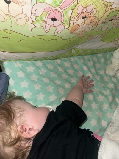 The media file shows a close-up, overhead view of a baby sleeping in a crib. The baby is lying on its side, with its head turned away from the camera and resting on a pillow or mattress. A pacifier is visible in the baby's mouth. The crib bedding has a light green background with white star patterns. To the left of the star-patterned bedding, there is another piece of bedding with a green and white checkered pattern and cartoon images of teddy bears and rabbits. A white, fluffy blanket is visible at the top of the frame. The lighting suggests it is indoors, possibly nighttime or a dimly lit room, creating a peaceful and cozy atmosphere. The focus is on the baby and its immediate surroundings, implying an intimate or personal setting like a nursery or bedroom. There is no visible text in the media file.
