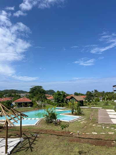 The image shows a resort-style swimming pool located in a tropical setting in Las Escobas del Venado, Panama, with coordinates 7.432 latitude, -80.188 longitude.  The pool has an irregular shape and is surrounded by a grassy area with a paved walkway leading to it. There are several palm trees and other tropical plants throughout the area.  A wooden structure, possibly a pergola, is located to the left of the pool, and a small wooden building is in the background. The sky is a clear blue with wispy clouds.  There are no people present.
