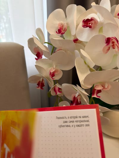 The image shows a close-up of white orchids with pink centers, partially obscuring a book with a red cover. The book's pages are visible, with text in Russian. The text reads, "Реальность, в которой мы живем, даже самая материальная, субъективна, и у каждого своя." This translates to, "The reality in which we live, even the most material, is subjective, and each person has their own." The background features a cream-colored armchair and a sheer white curtain. The lighting suggests an indoor setting, possibly a home or office. There is no indication of the city of Kazan, Russia, or any specific location.