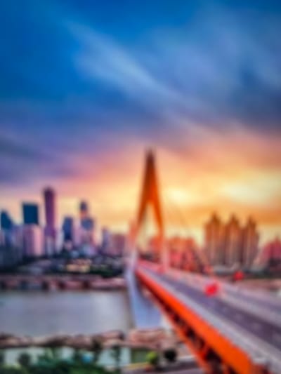 This is a heavily blurred, vertically oriented image depicting an urban landscape during either sunrise or sunset. The scene features a prominent, large bridge spanning across a body of water, with city buildings lining the distant banks.
The sky, occupying the upper and left portions of the image, displays a striking gradient. It transitions from a deep, rich blue in the far upper-left to a warm, vibrant orange and yellow on the right and towards the center, indicative of a transitional light period. This warm light illuminates parts of the cityscape in the background.
The bridge, stretching diagonally from the upper-right to the mid-left, is characterized by distinct orange or reddish structural elements, suggesting it might be a suspension or cable-stayed design. Due to the blur, specific details of the bridge's construction or any traffic on it are indistinguishable, though faint shapes might suggest vehicles or pedestrians.
Along the horizon, silhouetted against the bright, warm sky, are numerous indistinct city buildings, appearing as abstract blocks and vertical lines, suggesting a dense urban environment with skyscrapers. A body of water occupies the lower-right portion, reflecting the sky's colors.
No clear activity or specific events are discernible due to the extreme blur. There is no visible text. The overall impression is one of an atmospheric, almost painterly representation of a city at dawn or dusk, consistent with a major urban center like Yuzhong District, China, known for its dramatic landscapes and bridges.