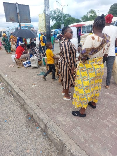 The scene depicts a busy urban street or bus terminal environment in Dar es-Salaam, Tanzania, under an overcast sky.
In the foreground, an asphalt road with scattered debris, including small colored plastic pieces, runs along the bottom left. It is bordered by a concrete curb separating it from a paved sidewalk constructed with reddish-brown interlocking bricks.
On the sidewalk, numerous individuals are present. A woman with dark skin stands prominently in the midground, facing left, wearing a long-sleeved, calf-length dress with vertical black and tan stripes and sunglasses. She wears light-colored flat sandals. To her right, another woman with dark skin stands with her back partially to the camera, facing right. She wears a cream-colored top with brown and white patterned trim and a long, bright yellow skirt with intricate blue, green, and orange patterns. She wears black flat shoes/sandals and has her hair styled in a bun. Between these two women and slightly to the left, a child wearing a bright yellow t-shirt and dark pants walks leftward.
Further left on the sidewalk, a man in a red t-shirt and blue baseball cap is seated on wooden crates under a black open umbrella. Another person in a plaid shirt is seated nearby on crates. A large concrete utility pole stands in the center midground, with a yellow sack and other materials attached. Various other individuals, including a woman in a green patterned dress on the far left and a man in a white t-shirt and red cap behind the yellow-skirted woman, are distributed across the sidewalk.
In the background, multiple buses, including light blue and white and red and white models, are visible, parked or in motion, indicating a public transport hub. Overhead streetlights and trees are also present in the distance. The woman in the striped dress exhibits a slight smile and is looking towards the right. No direct interactions between the prominent individuals are explicitly discernible.