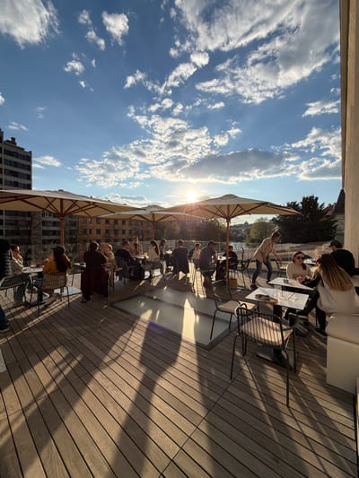 The photograph depicts an outdoor setting with people seated at tables under umbrellas on a wooden deck. The scene appears to be a rooftop terrace of a building in Ljubljana, Slovenia, based on the architectural style and the visible cityscape. The sun is shining brightly, casting long shadows across the deck. People are engaged in various activities such as talking, eating, and relaxing. There are buildings and trees visible in the background. The sky is blue with scattered clouds.