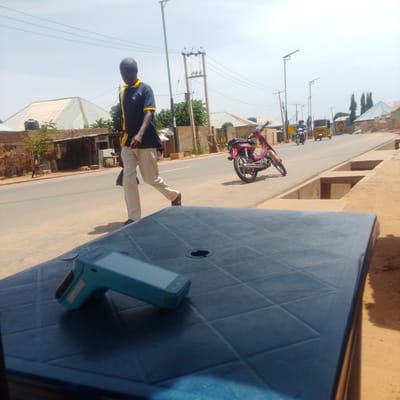 In this outdoor, daytime scene in Tempure, Nigeria, a man in a dark blue polo shirt with yellow trim and light-colored pants walks across a paved road. He is carrying a dark bag in his left hand. In the background, a red motorcycle is parked on the side of the road. Further down the road, a yellow auto-rickshaw and a motorcycle with riders are visible. The setting appears to be a street with buildings and shops lining one side, and a dry, sandy embankment on the other. The weather is clear and sunny, suggesting a warm day. In the foreground, a dark, textured surface, likely a table, is partially visible, with a light blue electronic device, possibly a scanner or a small tablet, placed on it. The scene captures a moment of everyday life in a Nigerian town.