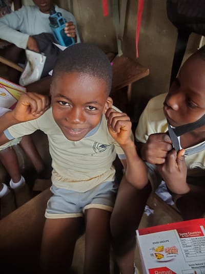 The image shows two children in what appears to be a classroom setting in Ukwu-Abwa, Nigeria. One child, positioned towards the bottom of the frame, is looking directly at the camera and smiling. This child is wearing a light yellow shirt with grey trim and grey shorts, indicating a school uniform. Part of a school logo is visible on the shirt. The child is seated at a wooden desk.
The second child is partially visible at the top of the frame, focused on adjusting a black strap, possibly part of a bag or uniform. This child is also wearing the same uniform shirt.
On the desk near the smiling child, there is an open textbook about nutrition and world currencies. The text and images indicate an educational context related to food groups and international finance.
In the lower-left corner of the image, part of a third person can be seen holding a blue object. The environment suggests a modestly furnished classroom interior.