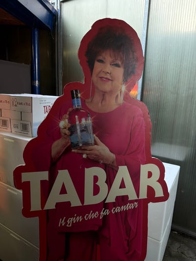The image shows a life-sized cardboard cutout of an Italian actress and singer, Patty Pravo, holding a bottle of Tabar London Dry Gin. The cutout is red and features the text "TABAR" in large white letters, with the tagline "Il gin che fa cantar" (The gin that makes you sing) below it. The cutout is positioned in front of stacked white boxes, one of which has labels indicating "6 bott. x 70 cl." and "PREMIUM GIN." The background appears to be a corrugated metal wall or partition. The scene suggests an outdoor or semi-outdoor commercial setting, possibly a market stall or a warehouse area, given the stacked boxes and industrial-looking background. There is no specific indication of the city of Milan, Italy, in the image itself; this information is provided externally. The lighting is moderate, with some shadowing.