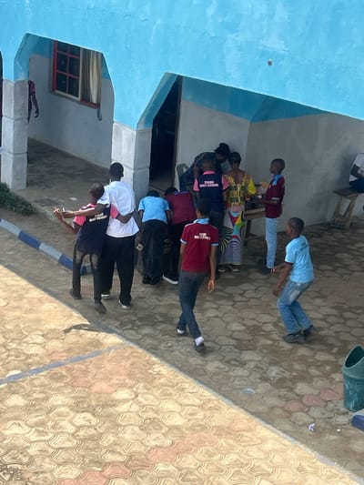 The image captures an outdoor gathering of approximately a dozen individuals, primarily young people and some adults, in what appears to be a paved courtyard or schoolyard in Ikorodu, Nigeria. The scene is brightly lit by natural sunlight, casting distinct shadows and suggesting it is midday or early afternoon on a clear, sunny day.
The subjects are mostly standing, some in various school uniforms. Several individuals wear dark blue polo shirts with white collars and sleeves, emblazoned with text that clearly reads "IKORODU HIGH SCHOOL." Others are in maroon polo shirts with light blue collars and similar text. Casual attire is also visible, including a person in a light blue t-shirt and jeans, another in a white polo shirt and black trousers, and an adult in a vibrant, patterned dress. One person in a black t-shirt also has white text, likely the school name.
To the left, a building with light blue painted walls and a red-framed window is partially visible. The ground is covered with decorative, interlocking pavers in shades of brown, grey, and reddish-pink. Scattered on the ground are small pieces of litter. In the upper right corner, a dark green bucket or bin is present. A low curb or barrier, striped in blue and white, runs across the lower middle section of the paved area. The people appear to be gathered informally, observing their surroundings or possibly awaiting an instruction, without any specific organized activity immediately apparent.