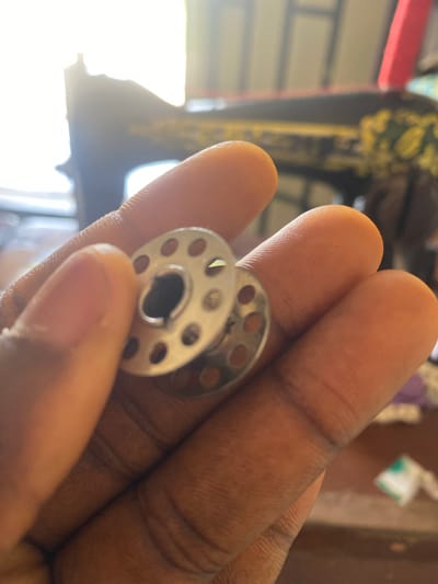 A close-up shot shows a person's hand holding two metal bobbins. One bobbin is in focus, and the other is slightly out of focus, underneath the first. The bobbins are circular with a central hole and several smaller holes around the perimeter. The hand is cupped, with fingers visible in the foreground. In the background, a sewing machine is partially visible, appearing dark with some yellow and green decorative elements. The scene is illuminated by a bright light source, possibly natural light, coming from the left, creating a strong backlight effect that silhouettes the sewing machine and parts of the hand. The overall environment suggests an indoor setting, possibly a home or workshop in Jauro Yinu, Nigeria.