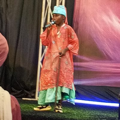 A full-body shot captures a person standing center-frame on a grassy stage in Sabon Tasha, Nigeria. The individual, adorned in a flowing, long orange garment with subtle grey patterns and a light blue headscarf, holds a microphone in their right hand and a walking stick in their left. The garment extends to the knees, revealing a layer of light blue fabric underneath. The person wears yellow sandals with black straps on their left foot and black and white shoes on their right. The background features a dark curtain on the left, a series of vertical light-colored poles, and a pink and purple draped fabric on the right. A bright blue light runs horizontally across the bottom right of the frame. The overall setting appears to be an indoor performance or presentation.