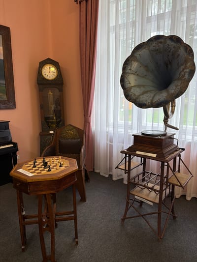 This media file depicts an indoor scene showcasing antique items. The main subjects are a gramophone with a large horn, a grandfather clock, a chessboard with pieces set up, and parts of a piano. The setting appears to be a room with a large window covered by sheer white curtains and a pink wall. There are also wooden chairs and a table. The floor is covered with dark grey carpet.
The gramophone is prominently featured in the upper left, its metallic horn pointing upwards and to the left. Next to it, a wooden cabinet with a record player is visible, supported by a rustic wooden stand with woven seats. A label is attached to the stand.
In the lower section, a grandfather clock with an ornate dial and pendulum is mounted on the pink wall. To its right, an octagonal wooden table holds a chessboard with black and white pieces in mid-game. Behind the chessboard, a leather-upholstered chair is visible. A portion of a piano keyboard is visible in the bottom center.
The lighting suggests daytime, with light streaming through the window. There are no people visible, and the scene is static, suggesting a display or a preserved historical room. The overall impression is one of nostalgia and historical preservation. No specific weather conditions can be inferred. The location context is Sarapul, Russia, but no explicit visual cues in the image directly confirm this. There is text on a label attached to the wooden stand of the gramophone, and also on a label on the chessboard, but the text is not legible enough to be transcribed.