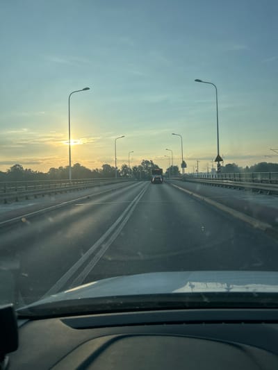 The image shows a view from inside a vehicle driving on a bridge over a body of water, likely during sunrise or sunset. The sun is visible low on the horizon, casting long shadows across the road and illuminating the sky with a soft, warm light. The bridge has multiple lanes of traffic and is lined with streetlights. Trees and vegetation can be seen on either side of the bridge, suggesting it is located in a suburban or natural area. A few other vehicles are visible on the bridge, including a car and a bus. The presence of overhead power lines in the distance indicates that the bridge is likely part of a larger transportation network. Without specific landmarks or identifying signs, it is difficult to pinpoint the exact location within Wrocław, Poland. However, the overall scene depicts a common urban or peri-urban landscape with infrastructure designed to facilitate transportation over water.