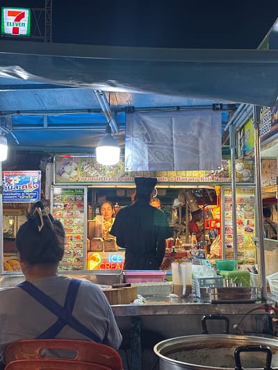 This nighttime street scene captures a bustling food stall in Koh Phangan, Thailand. The primary focus is on a vendor preparing food, with a woman's face visible as she appears to be serving. The stall is illuminated by bright overhead lights, casting a warm glow on the colorful displays of food items. Prominently featured are signs advertising "Coconut Ice-Cream," "Banana Roti Pancake," and various other treats, suggesting a sweet and possibly tropical offering.
Several individuals are present, including the vendor and what appear to be customers. The atmosphere seems lively, with the density of people and the bright lights indicative of a busy market or street food area. The stall itself is filled with a variety of packaged goods and ingredients, hinting at the culinary creations being made.
Visible text includes "ELEVEN," likely a brand reference, and Thai script on various signs, including what seems to be information about a government subsidy program ("คนละครึ่ง," "เป๋าตัง"). The overall impression is that of a vibrant, late-night food vending operation in a popular tourist destination. The time of day is clearly night due to the artificial lighting and the dark background. There are no indications of the weather.