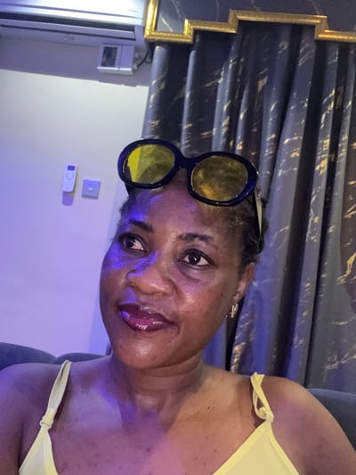 The image shows a close-up of a Black woman wearing yellow sunglasses on her head and a yellow top. She has dark skin, dark hair, and is wearing lip gloss. She is looking to her left. In the background, there is a dark blue curtain with gold accents and a white air conditioning unit mounted on a light-colored wall. There are also two white electrical outlets or switches on the wall. The lighting in the scene is predominantly purple. The image appears to be taken indoors, possibly in a residential setting in Ajara, Nigeria.