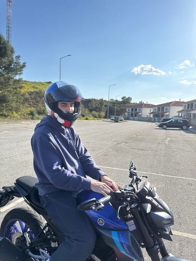 The image depicts a male individual seated on a blue and black Yamaha MT-125 motorcycle in an asphalt parking lot under a clear blue sky in Mirandela, Portugal. The subject wears a dark blue hoodie, dark blue jeans, and a multi-colored full-face motorcycle helmet with the visor partially open, revealing his face. His hands rest on the motorcycle's fuel tank. In the background to the left, a green hillside with some yellow vegetation is visible, topped by a red and white communication tower and a tree. To the right, a residential area features several two-story houses with light facades and red-tiled roofs, fronting a paved street where a black hatchback car is parked. Multiple streetlights are present in the background. White parking lines are visible on the asphalt surface. The scene is illuminated by bright sunlight.