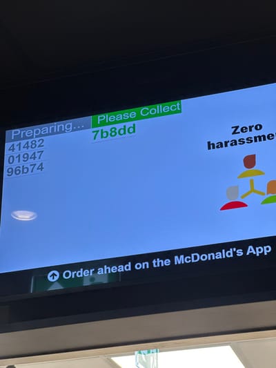 This image depicts a digital order status display screen, likely situated within a McDonald's restaurant in Chatham, United Kingdom. The screen exhibits a light blue background with various text and graphic elements. On the left side, under the grey banner "Preparing...", three alphanumeric order codes are listed vertically: "41482", "01947", and "96b74". To the right, a prominent green banner displays "Please Collect", below which the collection code "7b8dd" is visible. Further to the right, partially visible text reads "Zero harassment", accompanied by a graphic illustrating three stylized, interconnected human figures in brown, red, and yellow, signifying a network or group. Along the bottom edge of the screen, a dark bar features white text stating "Order ahead on the McDonald's App", preceded by a white upward arrow icon. A circular ceiling light fixture is reflected on the lower-left portion of the screen. The screen itself is enclosed by a dark frame, and a brightly lit interior space is visible beneath the display. No individuals are depicted within the frame.