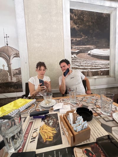 The image depicts two individuals, a woman and a man, seated at a table in an outdoor cafe setting, possibly in Cetinje, Montenegro, suggested by the presence of a Coca-Cola branded sugar holder and food items that may be typical of Balkan cuisine. The woman, with curly brown hair, wears a white t-shirt and is looking at her smartphone, which she holds in her hands. The man, with dark hair, is also looking at his smartphone, holding it to his ear as if in a call.
The table is set with various items including glasses of water, receipts, a yellow notepad, an ashtray, a sugar dispenser, and menus displaying food images, one of which shows french fries and meat. The background features a mural or large photograph depicting an outdoor amphitheater and a separate mural of a domed structure with a cross, possibly referencing historical or religious landmarks. The overall environment suggests a casual dining or break setting.