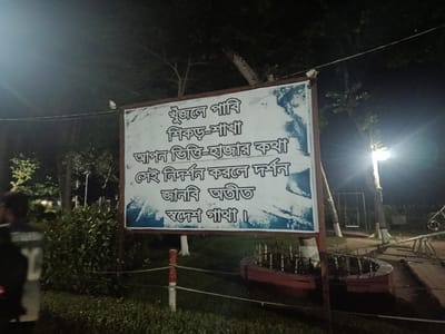 This is a nighttime, eye-level, medium shot of a sign in a park in Bagerhat, Bangladesh. The sign is white with dark text and features a blue and white image on the right side. The text appears to be in Bengali. The sign is supported by a brown metal frame and is illuminated by a bright overhead light.

The sign reads:
"খুঁজলে পাবি
শিকড়-শাখা
আপনি ভিত্তি-হাজার কথা
সেই নিদর্শন করলে দর্শন
জানবি অতীত
সদেশ গাথা।"

In English, this translates to:
"If you search, you will find
Roots-branches
A thousand foundation stories
When you see that evidence
You will know the past
The saga of the homeland."

To the left of the sign, a person is partially visible, walking away from the camera. They are wearing a dark jacket with the letter "Y" in white on the back.

The park is grassy with some bushes and trees visible in the background. There are also some playground elements, including a swing set and a seesaw, visible in the distance. The overall atmosphere is dark and somewhat eerie due to the nighttime setting and the bright light illuminating the sign. The ground is covered with grass and a red-colored border is visible around a circular planter in the foreground.