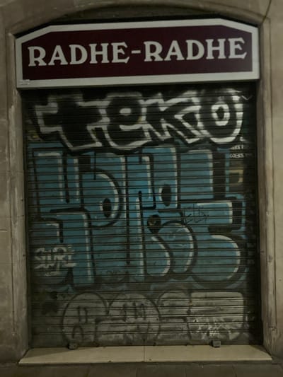 The image shows a closed metal shutter in Barcelona, Spain, covered in graffiti. Above the shutter, a sign with a dark red background and white text reads "RADHE-RADHE". The shutter itself is painted light blue and features large, white graffiti letters, possibly representing a tag. Below the main graffiti, there is another layer of faded graffiti in a darker color, and at the very bottom, what appears to be a stylized smiley face or abstract shape drawn in a light gray tone. The shutter is installed in a building with concrete pillars on either side. The lighting suggests it is nighttime.