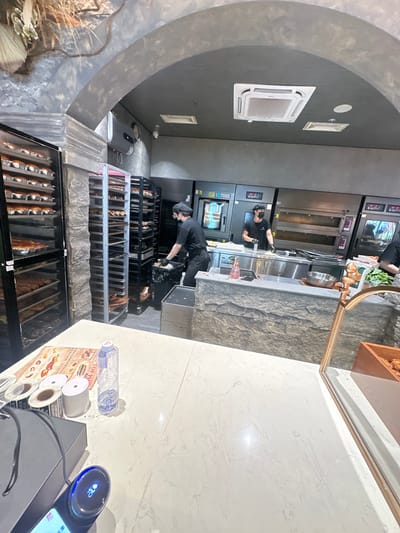 This image captures the interior of a commercial kitchen or bakery, likely located in Chongqing High-tech Zone, China. The scene shows multiple staff members working amidst industrial kitchen equipment and food display units.

In the mid-ground, two individuals in black uniforms and face masks are actively engaged in tasks. One person, positioned behind a stainless steel counter with sinks and various cooking apparatus, appears to be preparing or handling items. Another staff member is bending over a set of multi-tiered racks, possibly stocking or organizing packaged food products. The kitchen area features large ovens or baking equipment, some with digital displays showing numbers like "40:00" and "0.00."

The foreground on the right reveals a clean, white marble-like countertop. Various items are placed here, including a clear plastic water bottle (partially visible brand "Watsons"), multiple rolls of clear and white tape or labels (one showing "LANGKOU"), a flyer or menu, and a modern point-of-sale (POS) system or weighing scale with a blue illuminated display. The flyer has visible English text such as "TOP PI[CK]" and "RECOMMENDED SERIES," alongside unidentifiable Chinese characters. The POS system also displays numbers (e.g., "23") and what appears to be Chinese text.

On the left side of the image, a large, dark gray arched architectural feature frames the view. The overall lighting is bright and artificial, typical of a commercial establishment. Below the countertop and extending into the left side are numerous black and white racks filled with individually packaged baked goods or prepared food items, arranged neatly for display or storage. A pink bottle labeled "FREEZE" is also visible near the stainless steel counter.