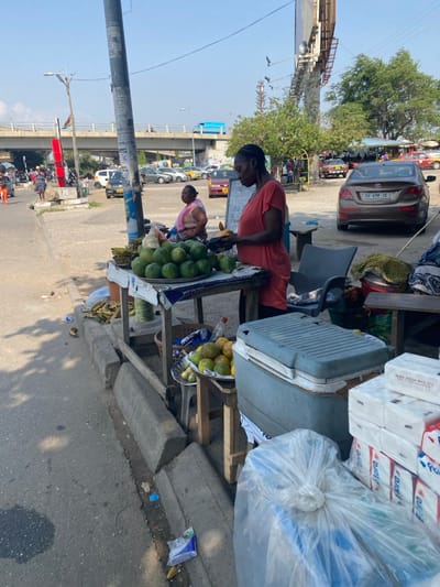The image portrays a street vending scene in Accra, Ghana, specifically near the Kwame Nkrumah Interchange. In the foreground, a dark-skinned woman wearing a reddish-orange dress stands behind a wooden fruit stall, actively preparing a light-colored fruit. The stall displays a metal tray filled with green-skinned fruits, likely oranges or grapefruits, alongside a bunch of bananas. Adjacent to this, a second wooden stall holds a smaller tray of yellow-orange fruits and what appear to be ears of corn. To the right, a light-blue cooler with a gray lid rests on the ground, next to stacks of white packaged goods, including "Flora" brand items and products from "WIDE MESH MFG LTD," along with a large clear plastic bag of indeterminate contents. A concrete curb separates the vendor area from an asphalt road, where scattered litter is visible.

In the mid-ground, a second dark-skinned woman in a pink top is seated on a wooden bench, observing the scene. The background reveals a busy urban environment: a brown Hyundai Accent (Ghanaian license plate GW 698-15) is parked on the right. Beyond the stalls, a parking area or unpaved lot contains multiple vehicles, including several taxis with yellow roofs. An elevated concrete overpass spans the upper background, with a light blue vehicle driving across it. A tall utility pole, covered with various notices, stands prominently in the center. A large, dark billboard structure is visible to the right of the utility pole, partially obscured by trees. The sky is clear but exhibits a slight haze.