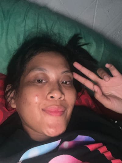 The image shows a close-up, selfie-style portrait of a young woman lying down. She is looking directly at the camera with a slight smile and has her right hand raised, forming a peace sign. Her hair is dark and pulled back loosely. She is wearing a dark colored t-shirt with a colorful, abstract graphic design. The background appears to be a green surface, possibly a blanket or pillow, and a red textured object, possibly another pillow. The lighting suggests it may be indoors or at night. The specific location in Kecamatan Pati, Indonesia, cannot be determined from the visual information provided in the image.