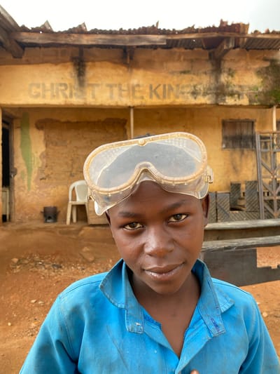 A slightly tilted, eye-level portrait shows a young person with dark skin, likely male, looking directly at the camera with a calm, engaging expression. He wears a light blue, long-sleeved collared shirt or work overall, which shows signs of wear and minor stains. A pair of clear plastic safety goggles with ventilation holes is pushed up onto his forehead.
The background features a rustic, weathered building with a yellowish-brown, textured wall. Faintly painted on the wall in light grey, the words "CHRIST THE KING" are partially visible, though faded and obscured. The building appears old, with some exposed structural elements, possibly wood or corrugated iron. A white plastic chair is visible in the mid-ground, near the building. The ground is a reddish-brown dirt or dry earth, suggesting an outdoor setting. In the upper background, indistinct metal structures, possibly scaffolding or part of a roof, can be seen. The bright, high-contrast lighting indicates it is daytime. The overall scene suggests a location in a rural or less developed urban area. The subject's attire and goggles hint at a work or vocational training context. The setting is Kuru Dungus, Nigeria.