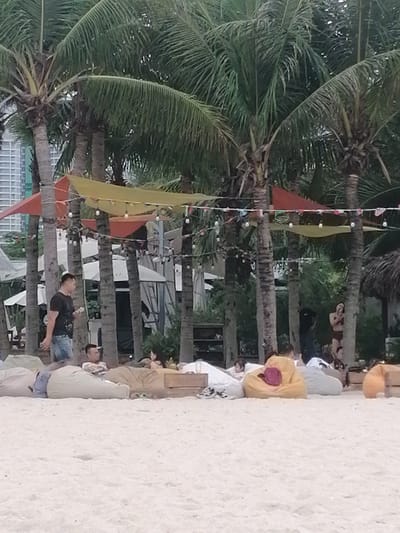 This image captures a relaxed scene on a sandy beach, likely in a resort or beach club setting in Bai Chay Ward, Vietnam. The foreground is dominated by a light-colored sandy beach. Scattered across the sand are numerous large, comfortable beanbags in various muted colors including beige, tan, yellow, gray, and orange, on which people are lounging.
Several individuals are visible. On the left, a man in a black t-shirt and denim shorts walks across the sand, holding a light-colored object. Further right, another man reclines on a beige beanbag, looking towards a woman sitting on an adjacent beanbag. In the background, under the shade of palm trees, other people are seen relaxing, with one person in a bikini near a thatched hut on the far right.
The midground and background are filled with numerous tall palm trees, their green fronds creating a canopy. Between the palm tree trunks, colorful fabric canopies in yellow, orange, and red are strung up, providing shade or decoration. Strings of decorative bare-bulb lights are also visible, draped between the trees and canopies, suggesting the scene might transition into evening. Behind the palm trees on the left, modern, multi-story buildings can be seen, indicating an urban or developed coastal area. White tents or large umbrellas are also visible further back, contributing to the resort atmosphere.
The time of day appears to be late afternoon or early evening, given the soft, diffused light, and the weather seems pleasant and calm. The overall mood is one of leisure and relaxation. There is no visible text in the image.