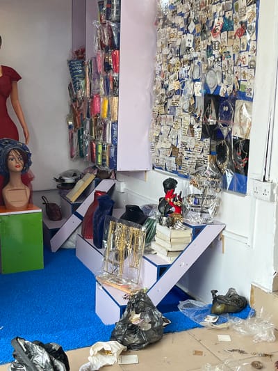 This image shows the interior of a shop in Ado, Nigeria. The walls are painted white, and shelves filled with merchandise line them. One shelf is packed with jewelry, mostly earrings and rings, displayed on small cards. Below this are shelves filled with colorful purses and clutches. A mannequin head adorned with a blue head wrap and a mannequin torso dressed in a red dress are placed at the bottom of the frame.

The shop's layout is organized with displays placed in a hexagonal format, covered in bright blue carpeting. Merchandise, including jewelry and accessories, are arranged on the display pieces. The overall impression is a small retail space focused on fashion accessories. The lighting is bright, giving good visibility to the products.