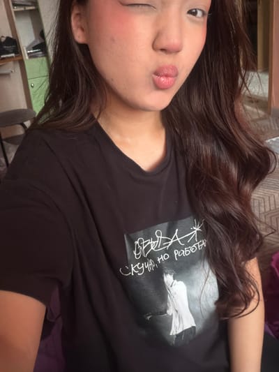 The image is a selfie of a young woman lying down. She has long, dark, wavy hair that falls around her face and over her shoulder. Her face is turned towards the camera, and she is looking slightly to the side with one eye closed. She is wearing makeup, including blush on her cheeks and pink lipstick on her lips. She is wearing a black t-shirt with a graphic design on it. The design includes a black and white image of a person in a dress and what appears to be handwritten text in white. The text is difficult to read clearly but appears to be in Cyrillic script.
The background is out of focus, but hints of a domestic setting are visible. To the left, there are shelves with some items on them, including what looks like a green cabinet. The lighting suggests it is daytime, and the overall mood is casual and relaxed. The image is taken from a close-up perspective, focusing on the woman's face and upper body. The orientation of the image is rotated.