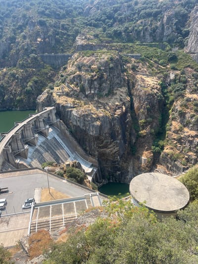 The image presents an elevated, wide-angle view of a large concrete arch-gravity dam situated within a deep, rocky gorge near Miranda do Douro, Portugal. The dam's downstream face, characterized by a curved, stepped concrete structure with visible buttresses, descends into a dark green plunge pool at its base. An expansive body of dark green water, consistent with a reservoir, is visible upstream of the dam, contained by steep, arid rock walls.
The surrounding environment consists of rugged, irregular rock formations and cliffs, primarily covered with sparse, dry, yellowish vegetation, interspersed with patches of denser green foliage, particularly on the upper slopes and along crevices.
In the foreground, a paved asphalt road or viewing area is visible, with a section featuring several parked passenger vehicles, including at least one white and two grey/silver cars. A single dark-colored utility pole stands on this paved surface. Below this road, a series of concrete steps or terraced platforms descend. To the right, partially obscured by green foliage, is a large, flat, circular concrete structure, consistent with an observation platform or a component of the dam's infrastructure. Distant in the background, horizontal lines on the steep rock faces suggest other constructed pathways or roads traversing the mountainside. The lighting indicates clear daylight conditions. No individuals are present in the scene.