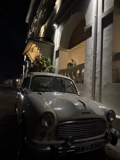 The image shows a white vintage car parked at night, possibly in a covered area or under an awning. The car is oriented at an angle, with its front grille and headlights prominently featured. The car's design suggests it might be from the mid-20th century, with a classic chrome grille and rounded headlights.
The setting appears to be an architectural structure with modern elements. Overhead, there are beams and ceiling structures, some of which are illuminated by warm lighting. In the background, through an opening, a more ornate architectural style is visible, possibly featuring columns and decorative elements. There is also some greenery, appearing to be palm-like plants, suggesting a tropical location.
The lighting is dim, indicating it is nighttime. The car's paintwork reflects the available light, giving it a polished appearance. No people are visible in the frame. The overall mood is quiet and possibly a bit mysterious due to the low light and the imposing presence of the vintage vehicle.
A license plate is partially visible on the front of the car, with some characters readable, suggesting it is **DLF-3304**. The location context of Galle, Sri Lanka, further supports the presence of tropical foliage and architectural styles often found in that region.
