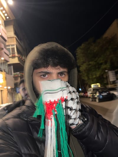 A person wearing a black puffer jacket and a hooded sweatshirt is partially covered by a kaffiyeh, with the bottom half featuring green, white, and red stripes with tassels. Only the person's eyes and forehead are visible. The background shows a dark sky, some foliage, and indistinct buildings illuminated by streetlights at night. The image is rotated, causing the background to appear vertically along the left side.