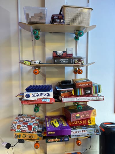 A vertical, multi-tiered shelf constructed from skateboard decks displays a variety of board games and other items. The shelves are supported by clear plastic poles, and the skateboard trucks and wheels are visible as decorative elements.
On the top shelf, a clear plastic bin contains wooden blocks, and a beige laundry basket sits next to it. Below that, a skateboard deck holds a few more items, including a small box with a skull design and a box labeled "Forbidden Desert".
The next level down features another skateboard deck with a small can and some game boxes. A larger game box labeled "SEQUENCE" is prominent on the shelf below, stacked with "Rummikub Classic".
Further down, a shelf holds a stack of games including "BOOGLE", "WAVELENGTH", and "CODENAMES".
The bottom shelves showcase more board games. One shelf has "AZUL" and "Dixit", while another holds "The Settlers of Catan" and "Ticket to Ride: Europe". A speaker is visible to the right of the lowest shelf.
The scene appears to be indoors, with a white wall in the background. There are no people visible in the image. The arrangement suggests a personal collection of board games organized on custom shelving. The presence of a speaker might indicate a recreational space. The image does not contain any identifiable landmarks or context to place it in Chiang Mai City Municipality, Thailand.