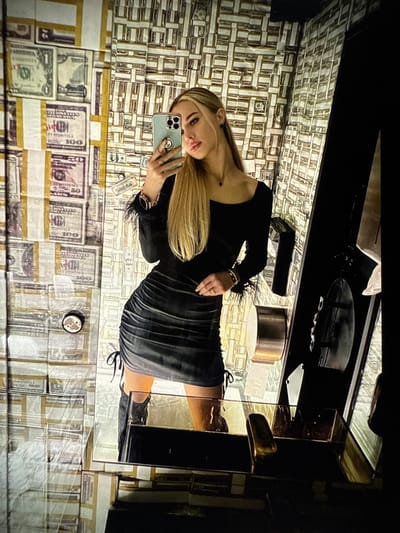 A blonde woman is taking a selfie in a restroom, reflected in a mirror. The walls of the restroom are decorated with a pattern of US dollar bills. The woman is wearing a black, long-sleeved dress with feather accents on the sleeves and a black skirt with a ruched effect and tie details. She is also wearing black boots. The restroom appears to be modern and luxurious, with gold accents on the fixtures. The overall impression is one of wealth and opulence. The image does not contain information to determine the location in the city of Denver, United States.