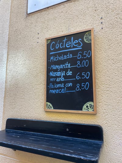 A wooden framed chalkboard menu lists cocktails and their prices in Spanish. The menu is mounted on a cream-colored wall, adjacent to a black wooden shelf. The menu is in Spanish, with a black background and handwritten white text, and includes a drawing of a slice of lemon. The shelf has a slight curve to it and a worn, dark black finish. The image was taken in Segovia, Spain, at the coordinates 40.95, -4.125.