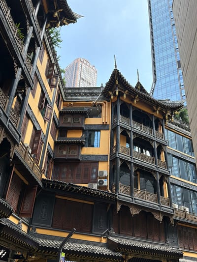 The image shows a view of traditional Chinese architecture against a backdrop of modern skyscrapers in Yuzhong District, China. The foreground is dominated by a multi-story building with a yellow facade, dark wooden balconies with intricate lattice work, and tiled roofs. Numerous wooden windows, some open, are visible. The building features ornate carvings and decorative elements. To the right and behind this structure, a sleek, modern glass skyscraper rises, its facade reflecting the sky and surrounding buildings. Further in the background, another tall building with a lighter facade and numerous windows is visible. The sky is clear and bright blue. There are no people visible in the image. The scene captures a juxtaposition of old and new architectural styles within an urban environment.