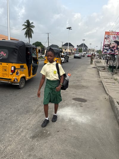 A full shot presents a young person with braided hair, wearing a tan school uniform with a green emblem and dark green shorts, standing in a street. The individual has a black bag slung over their shoulder and is wearing black shoes with white socks. They are positioned in the center of the street, with their left arm extended downwards.

The background shows a typical street in Ado, Nigeria. Yellow "keke" tricycles are parked alongside the road, one of which is in the immediate foreground. Beyond the tricycles, cars and pedestrian traffic are visible, moving along the street. Utility poles with electrical wires run along the street on both sides. A palm tree is visible on the left side, and a billboard advertising a hair and nail salon can be seen atop buildings on the right. The sky is overcast. The road surface is paved and appears to be concrete.